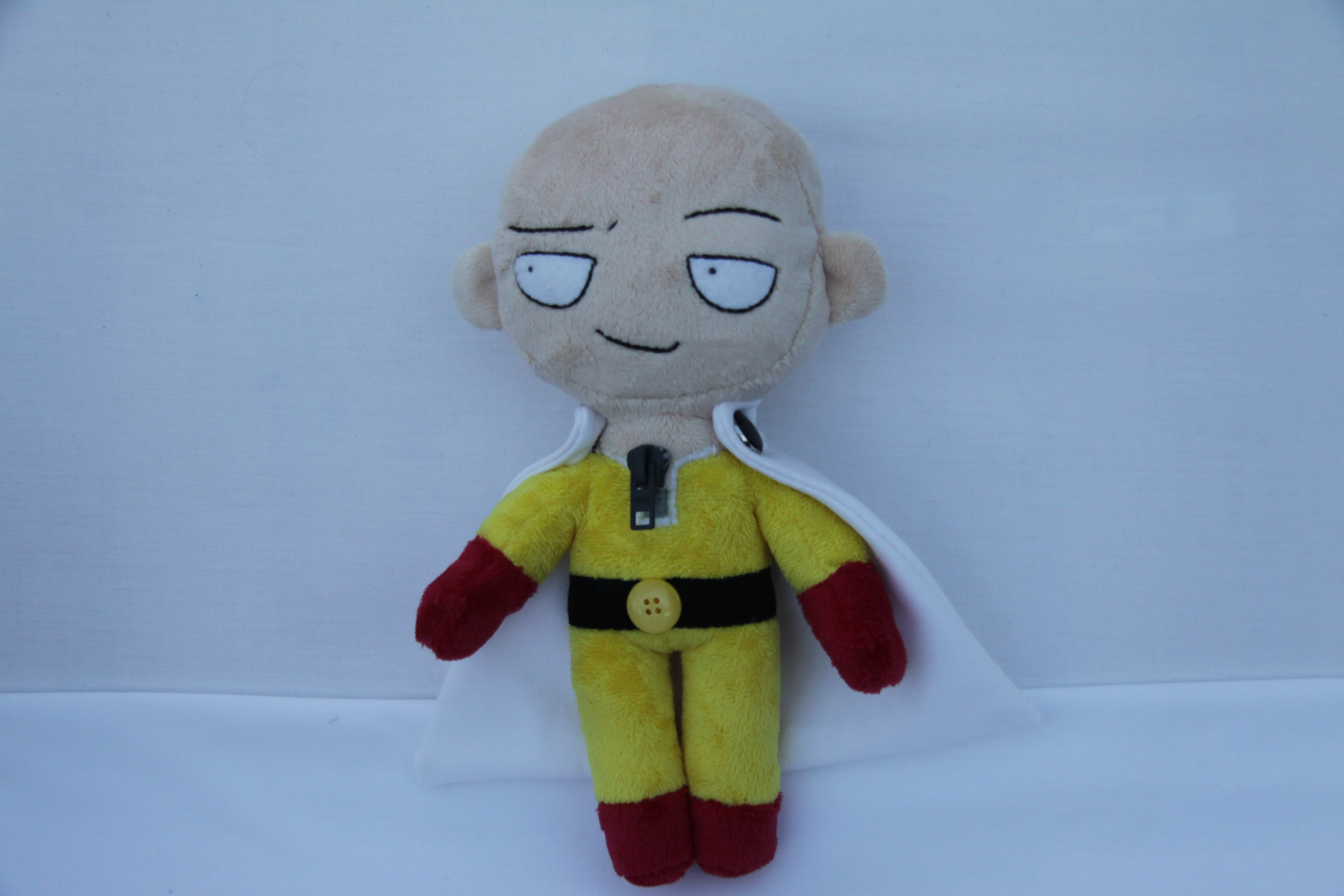 a plush of Saitama, a bald man wearing a yellow jumpsuit with a white cape held by black buttons. The jumpsuit is accented by a black belt with a yellow button buckle, and a grey front zipper.